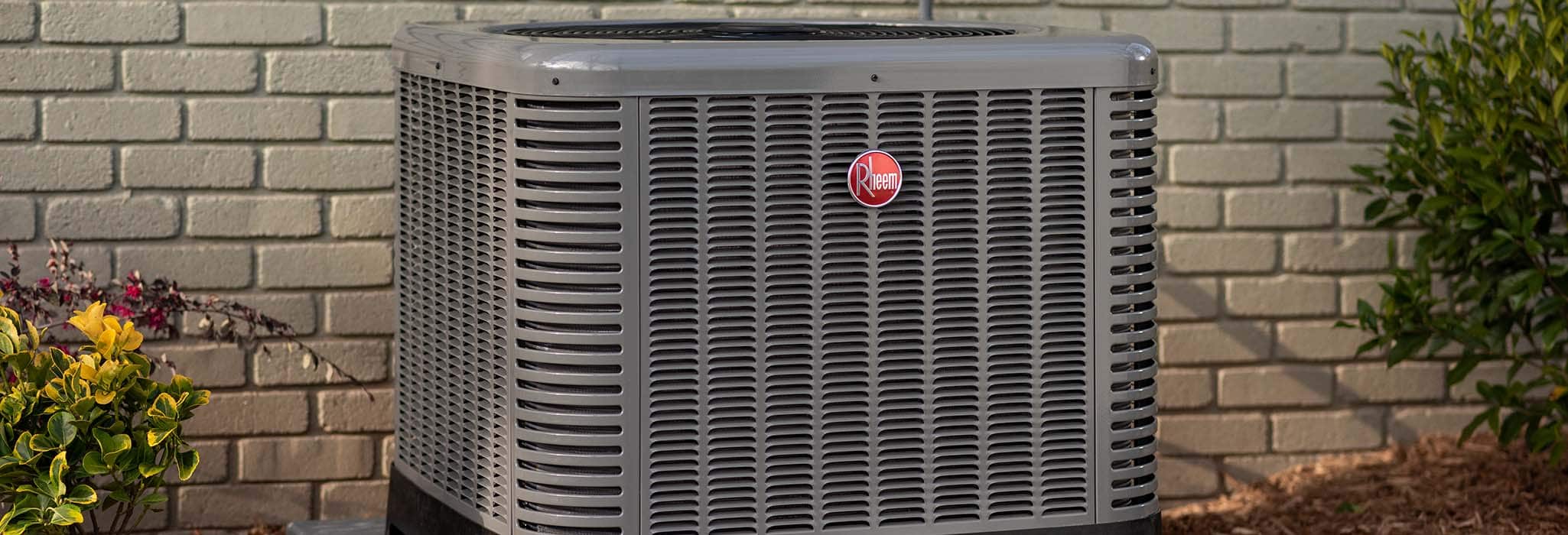 Rheem Air Conditioner unit sitting outside of a house against a brick wall.